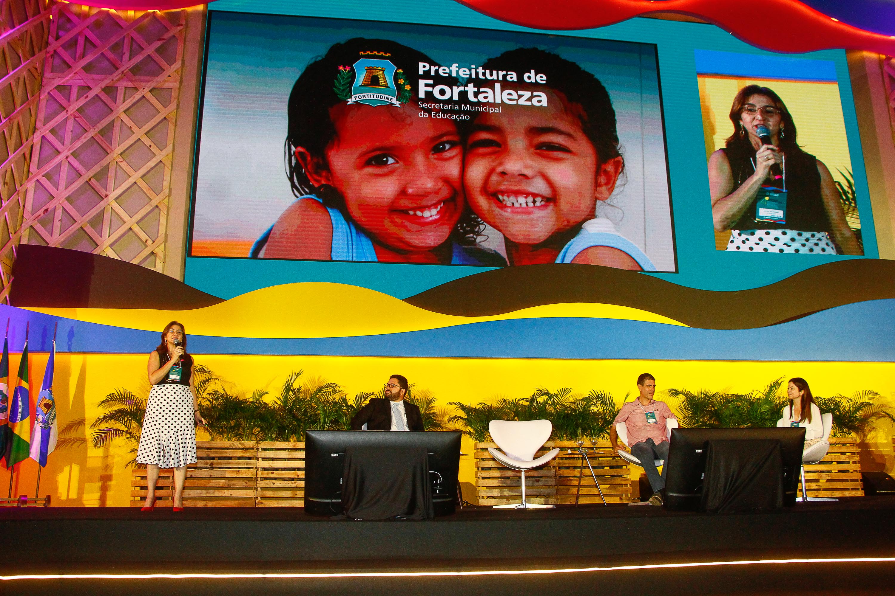 secretária Dalila Saldanha falando ao microfone em cima de palco, dois homens e uma mulher sentados atrás assistindo e painel ao fundo com foto de crianças